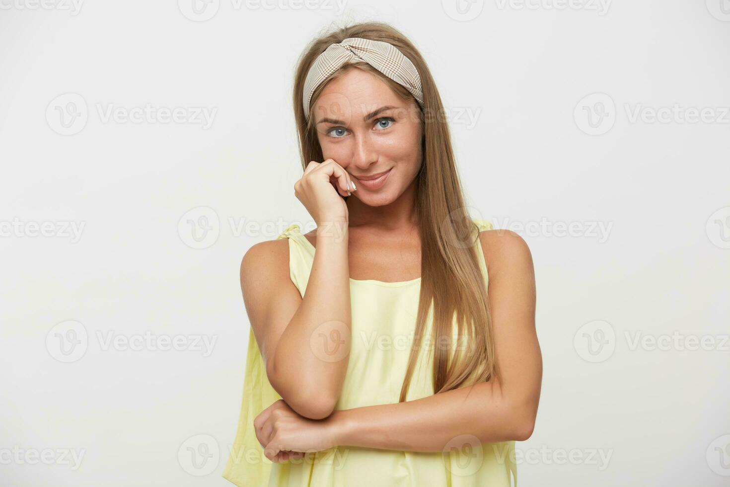 Romantic young attractive blonde woman with natural makeup looking positively at camera and smiling gently, touching soft her cheek with hand while posing over white background photo