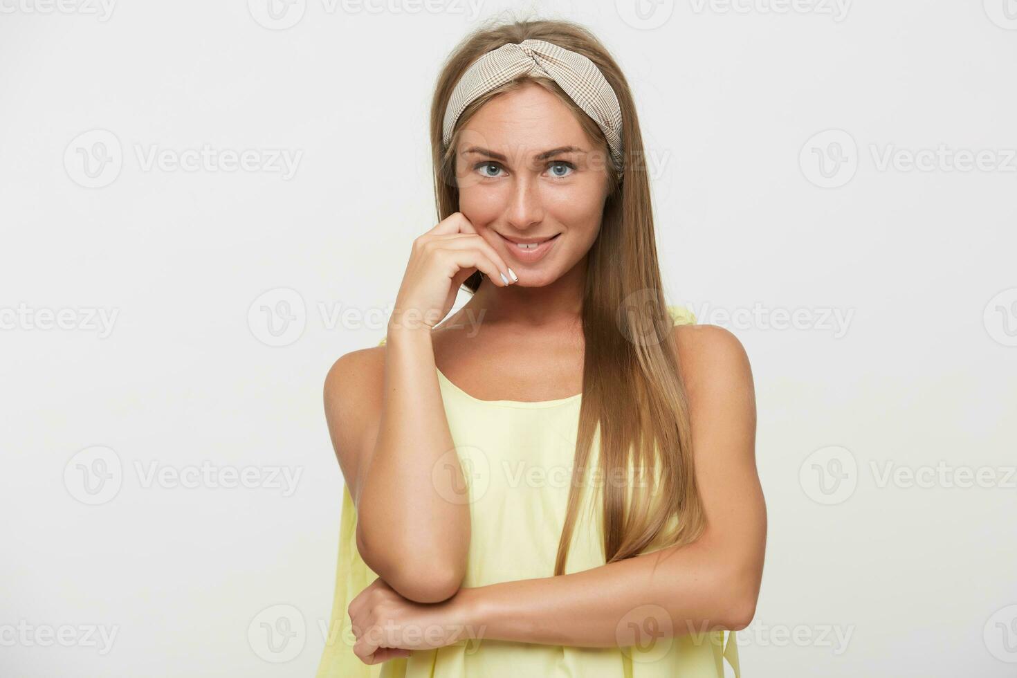 Indoor photo of beautiful positive young blonde lady with natural makeup touching gently her face with raised hand and smiling pleasantly, standing against white background