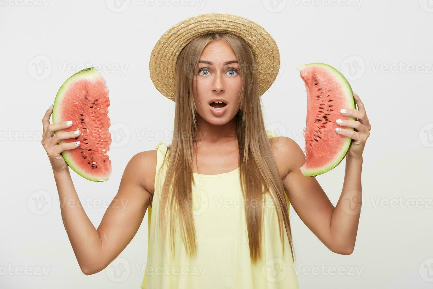 Surprised young lovely long haired blonde lady keeping her mouth wide opened while looking amazedly at camera, keeping pieces of watermelon in raised hands while posing over white background photo