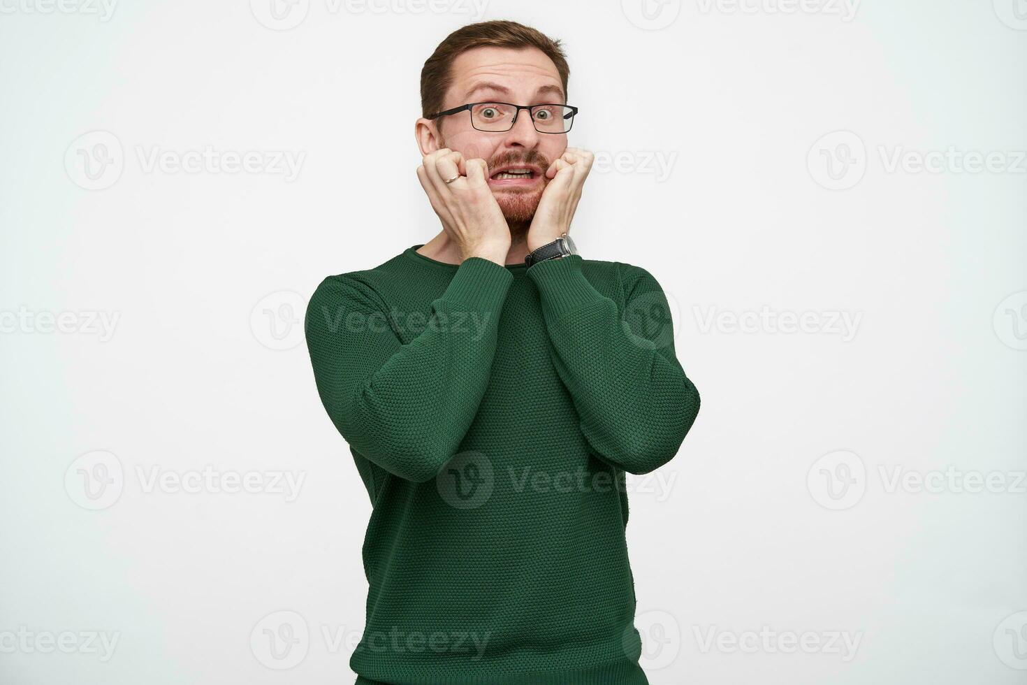 Indoor shot of scared young brunette bearded man in eyewear holding raised hands on his cheeks and crooking face afraid, standing against white background in casual clothes photo