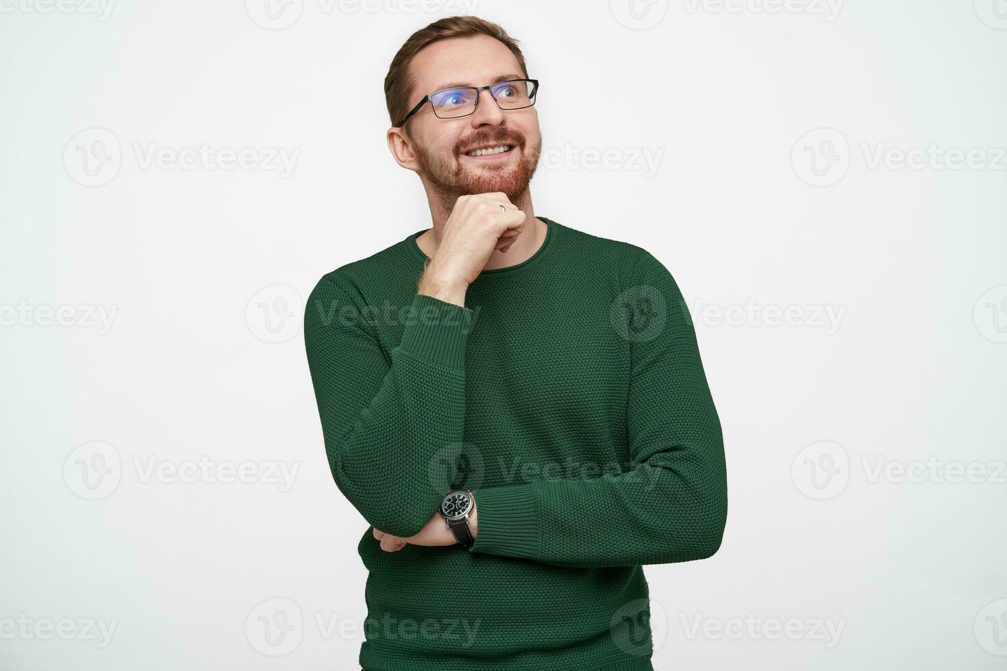 Cheerful young bearded male with brown short hair wearing glasses while posing over white background, raising hand to his chin and smiling positively while looking upwards photo