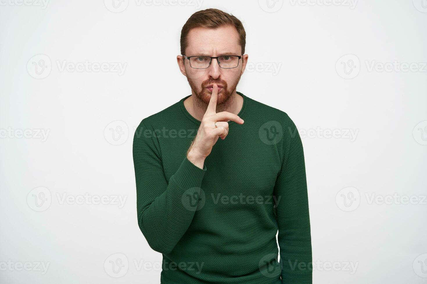 interior foto de joven morena barbado chico en lentes levantamiento mano con Cállate gesto mientras posando terminado blanco fondo, ceñudo Cejas y mirando seriamente a cámara