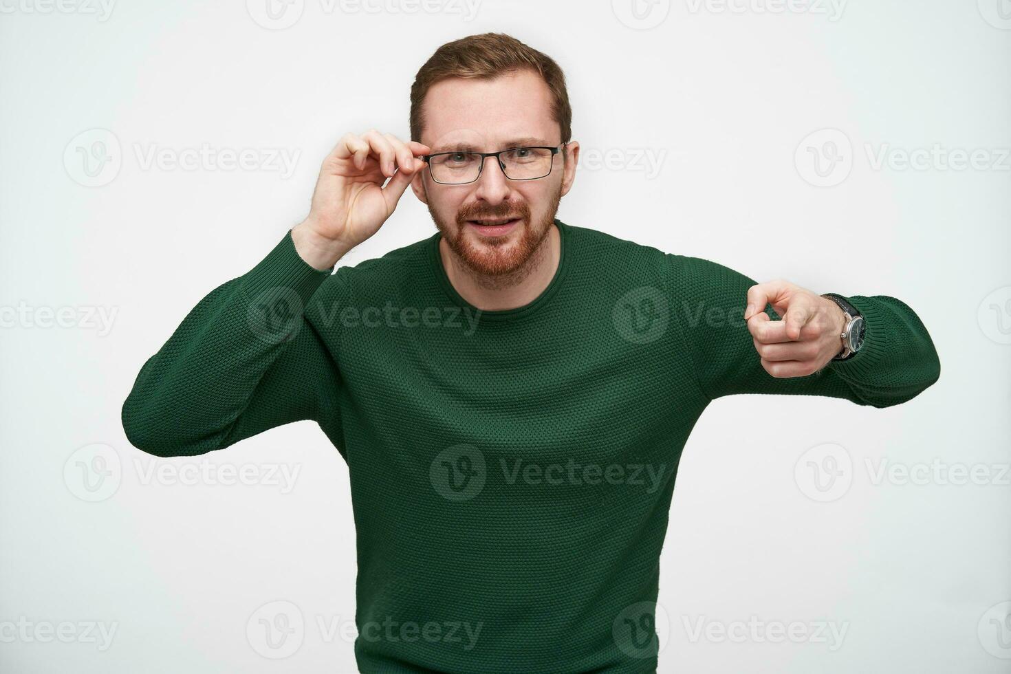 Studio shot of young puzzled brunette bearded guy holding his glasses with raised hand and pointing to camera with forefinger, frowning face while posing over white background photo