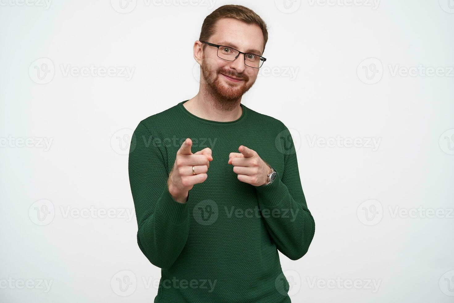 Positive young lovely brunette bearded man with short haircut pointing to camera with raised forefingers and smiling nicely, wearing green sweater and glasses over white background photo
