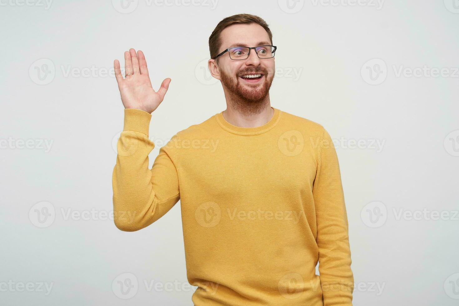 Shot of cheerful young handsome brown haired bearded guy in eyewear raising palm in hello gesture while looking positively aside with sincere smile, posing over white background in mustard pullover photo