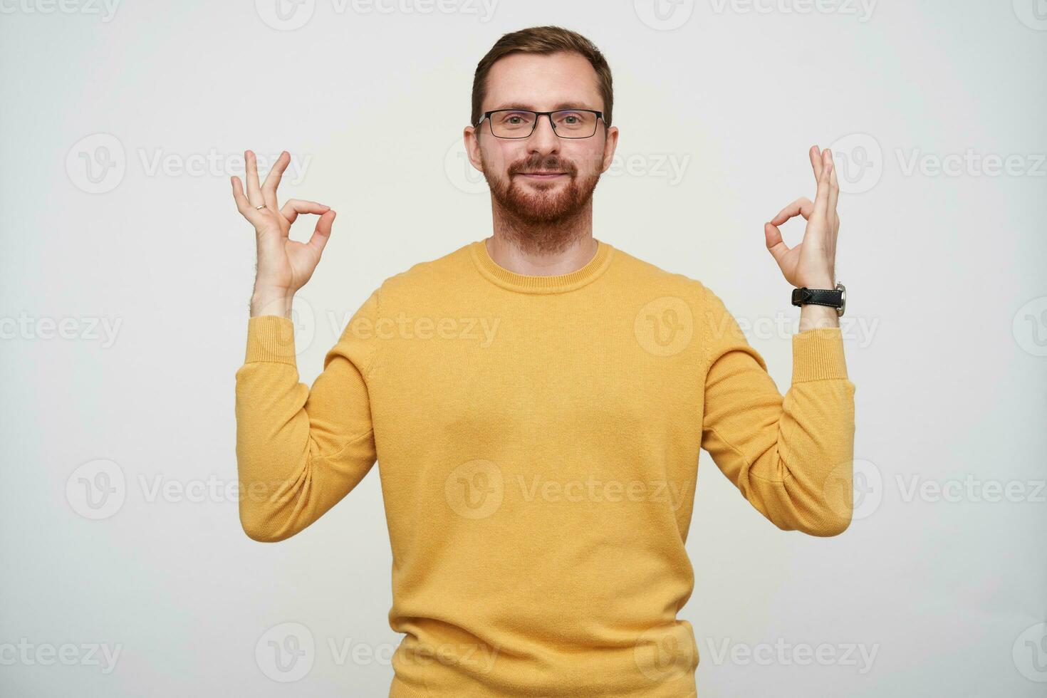 Indoor shot of attractive young bearded man in glasses with brown short hair folding fingers in mudra sign while posing over white background, looking at camera positively with calm face photo