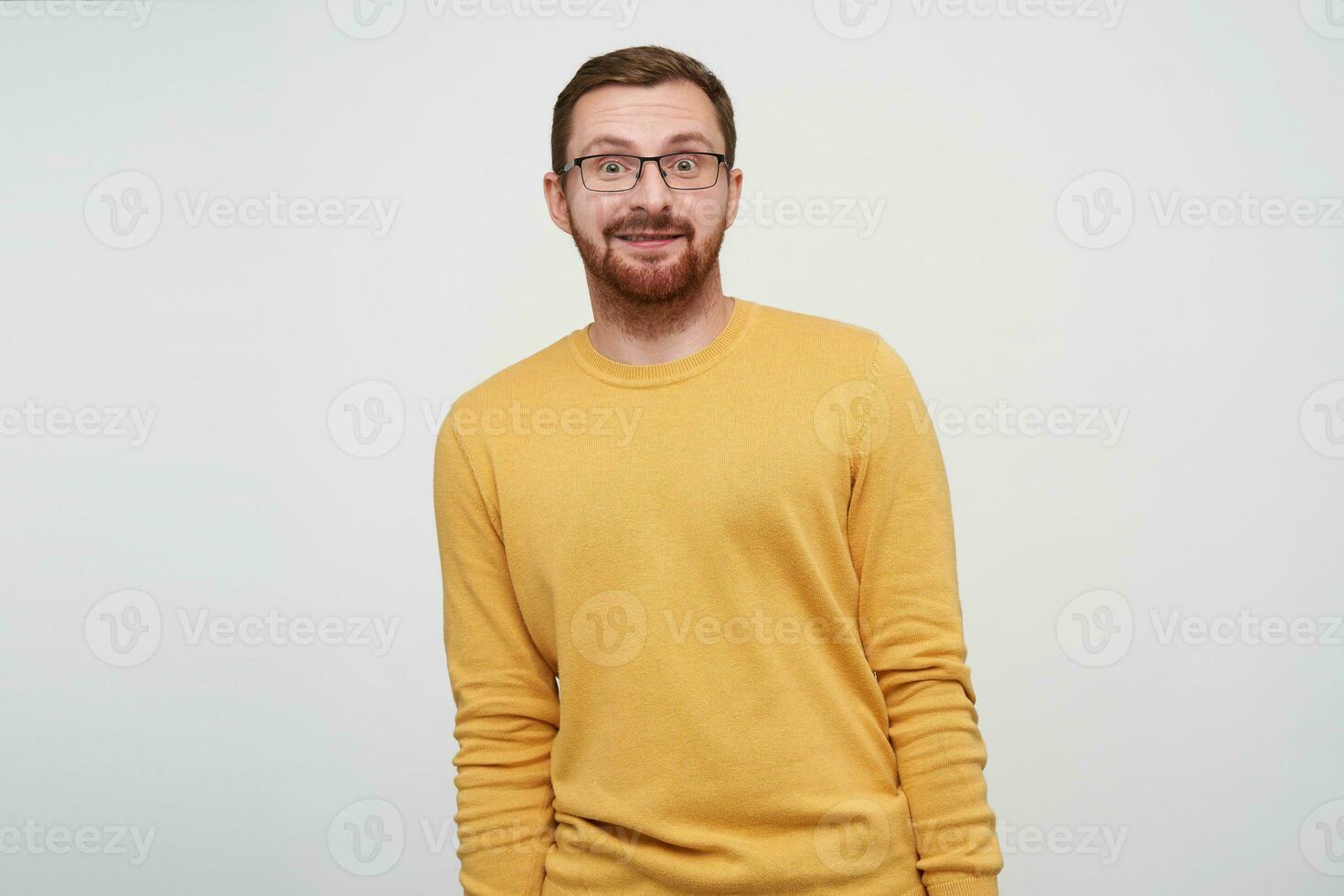 Surprised young brown haired bearded man wearing glasses while standing over white background with hands down, rounding eyes and raising amazedly eyebrows photo