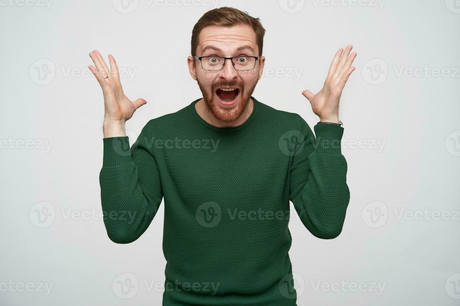 Overjoyed young pretty brunette bearded male in glasses raising happily hands and looking at camera with wide eyes and mouth opened, standing against white background photo