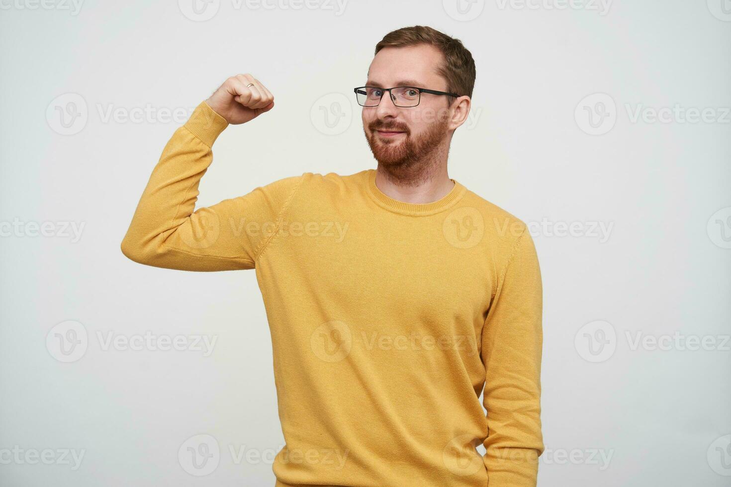 Portrait of cheerful young bearded male with brown short hair looking at camera self-confidently while demonstrating power in raised hand, standing against white background in casual clothes photo
