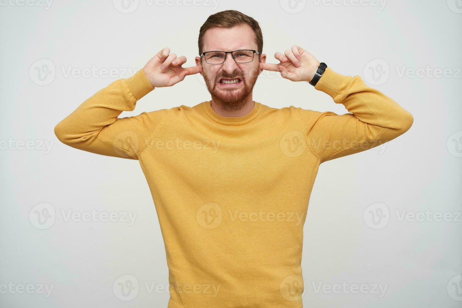 Dissatisfied young pretty bearded brunette man in glasses having short haircut, closing his eyes and frowning face while avoiding loud sounds, isolated over white background photo