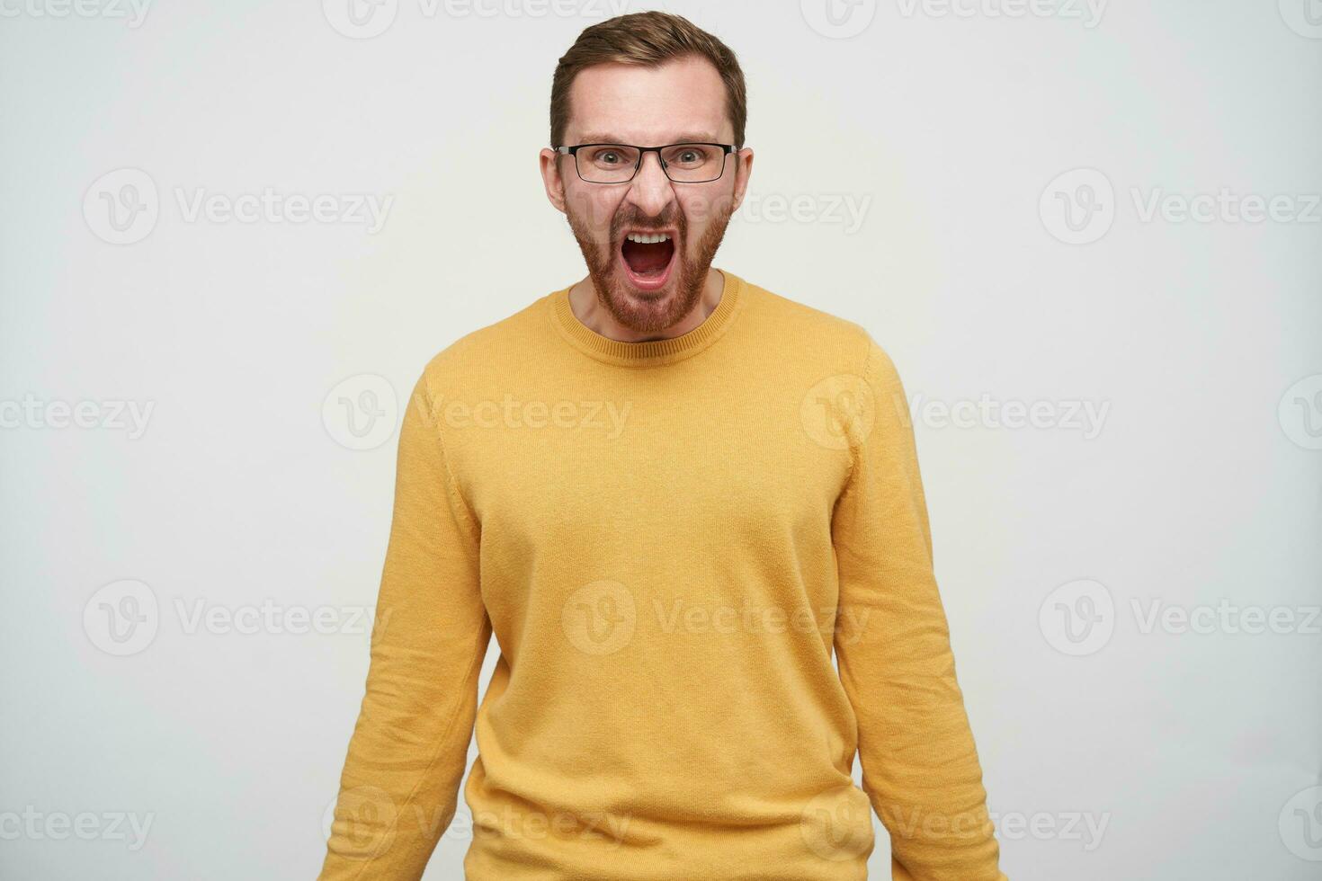 Furious young bearded male with brown short hair looking at camera and screaming violently with wide mouth opened, dressed in mustard pullover while standing over white background photo