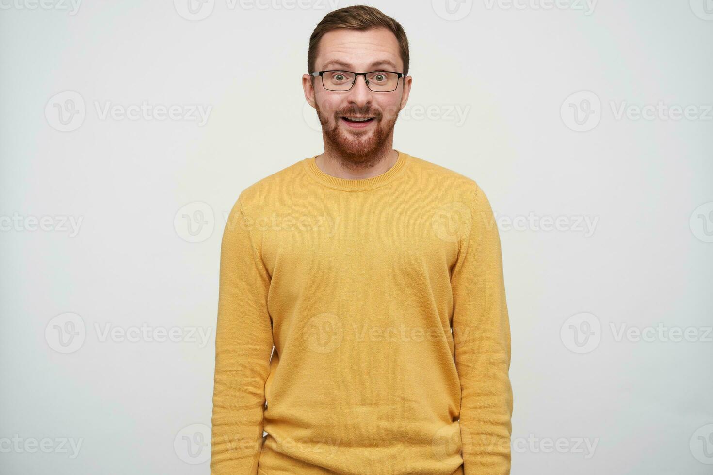 Suprised young brown haired pretty guy with beard wearing casual pullover and glasses while standing over white background with hands down, looking surprisedly at camera photo