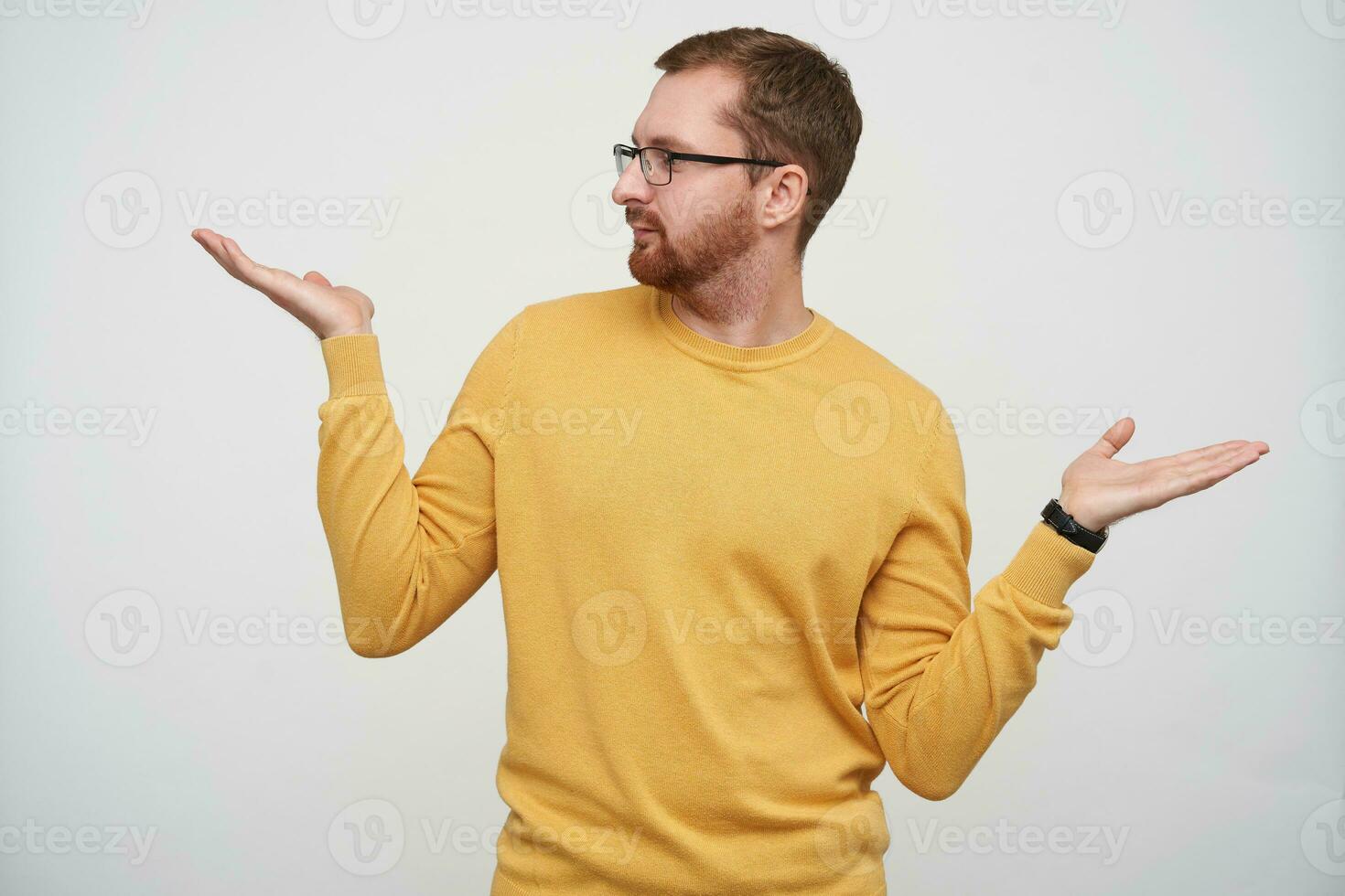 Positive young hansome brunette male with beard and short haircut wearing glasses and mustard pullover while standing over white background with raised palms photo