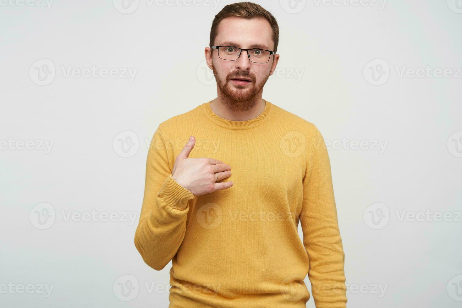 retrato de joven morena barbado chico en mostaza suéter vistiendo lentes mientras posando terminado blanco fondo, mirando confusamente a cámara y señalando en él mismo con elevado palma foto
