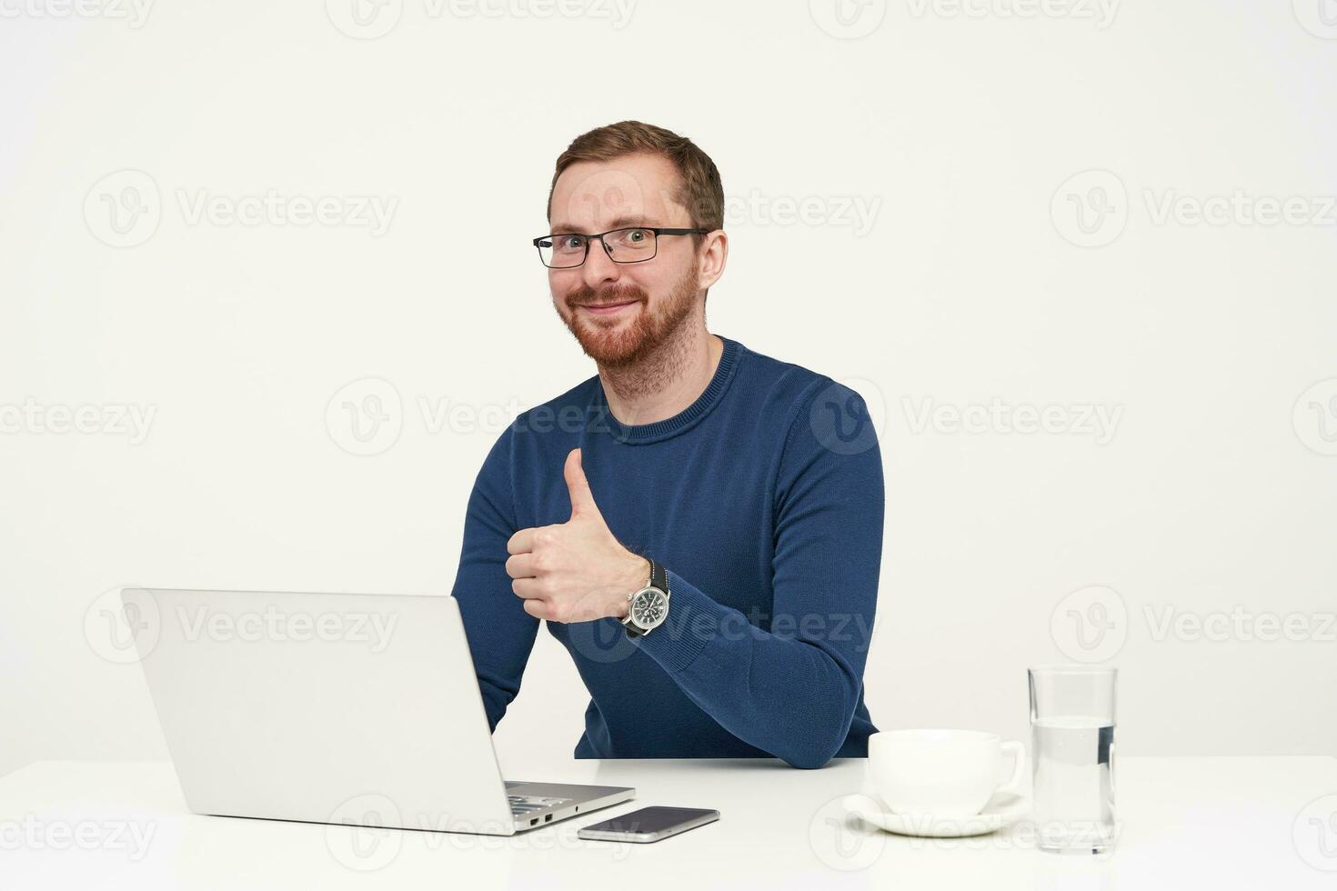 Studio shot of young positive fair-haired male in glasses showing raised thumb and smiling slightly at camera while sitting at table over white background photo