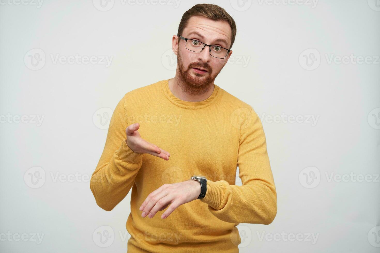 Dissatisfied young brown haired bearded guy in eyewear pointing indignantly on his with wristlet watch, being discontented that someone is late, isolated over white background photo