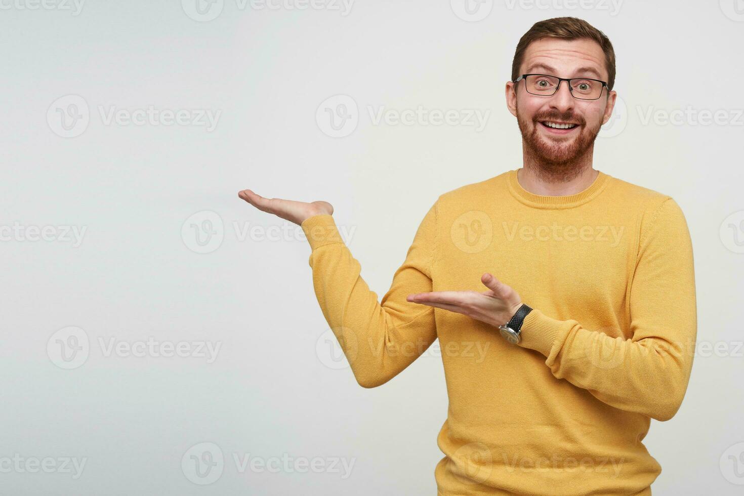 Indoor photo of joyful pretty young brunette bearded man with short haircut pointing aside with palms and raising happily eyebrows while looking at camera, isolated over white background