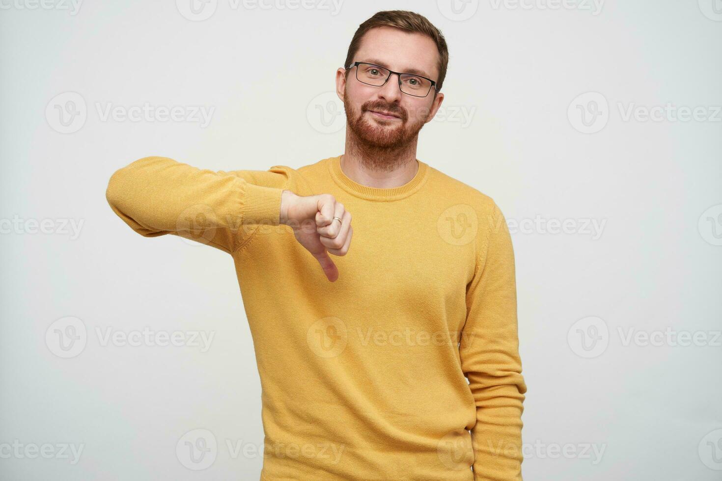 Studio photo of handsome young brunette bearded guy in glasses showing down with thumb and looking at camera with folded lips, isolated over white background