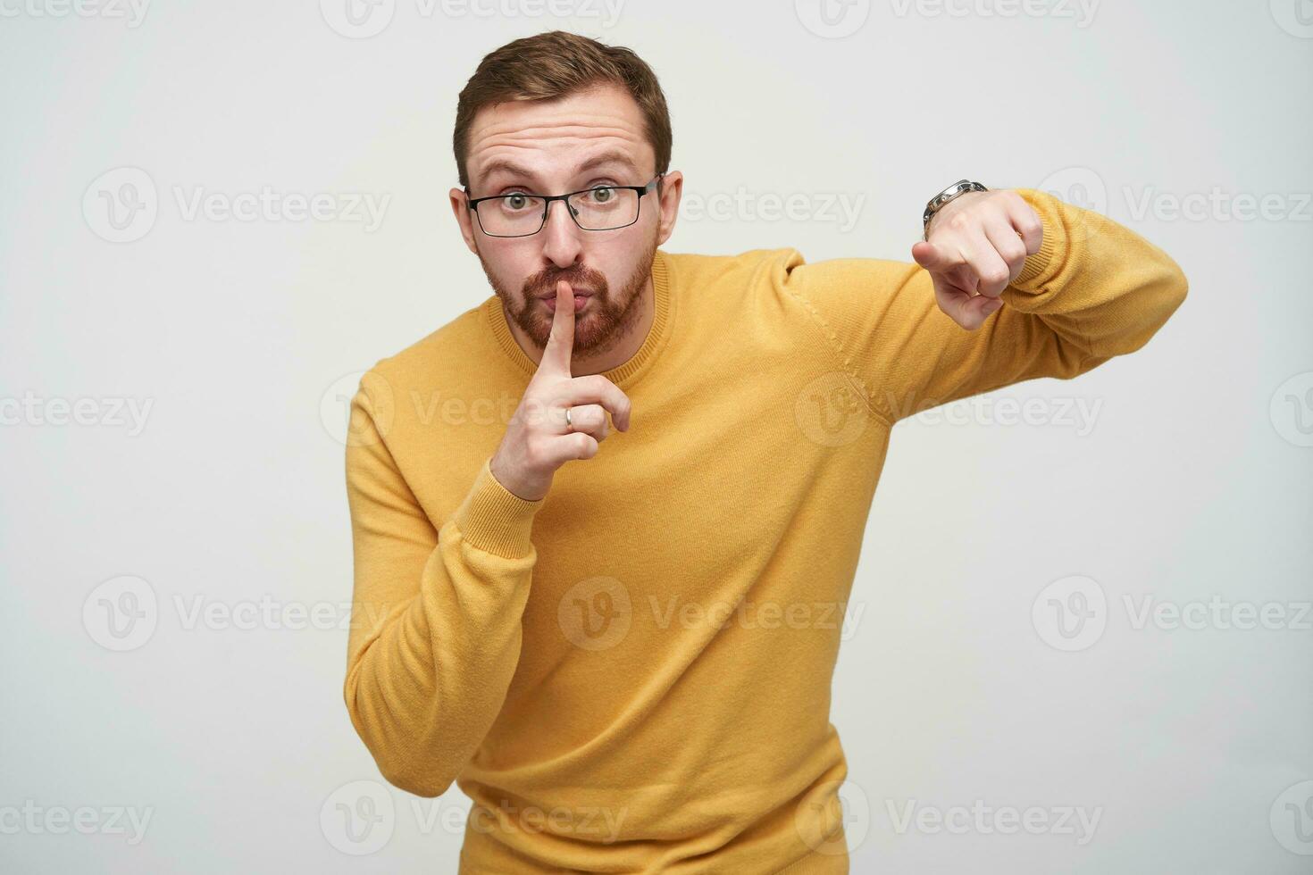 Serious young brunette short haired man with beard pointing on camera and keeping forefinger on his lips, asking to keep silence in late time, standing against white background photo