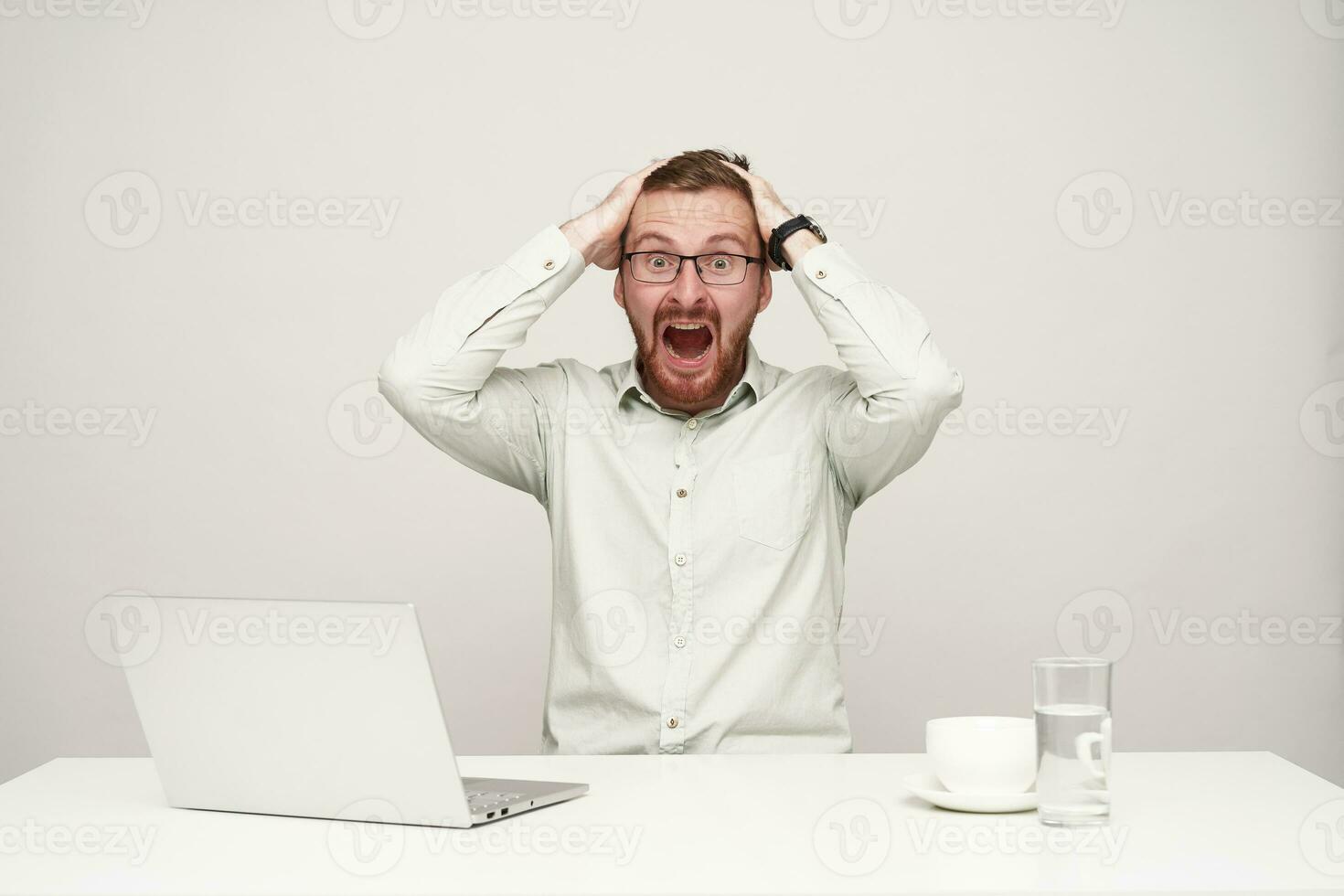 Studio photo of dazed young fair-haired male in formal clothes holding his head with raised hands and looking amazedly at camera with wide mouth opened, sitting over white background