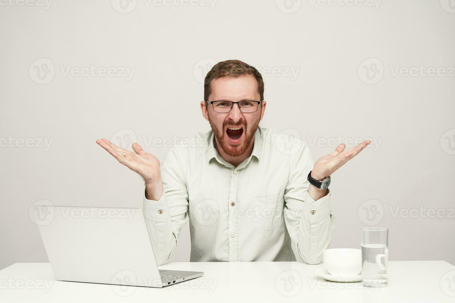 Stressed young bearded fair-haired male in glasses raising emotionally his palms while screaming with wide mouth opened, isolated over white background photo