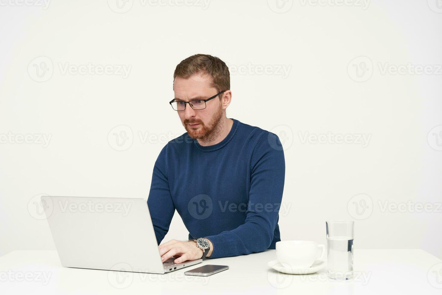 Indoor shot of young concentrated bearded guy in glasses looking seriously on screen of his laptop while working and keeping hands on keyboard, isolated over white background photo