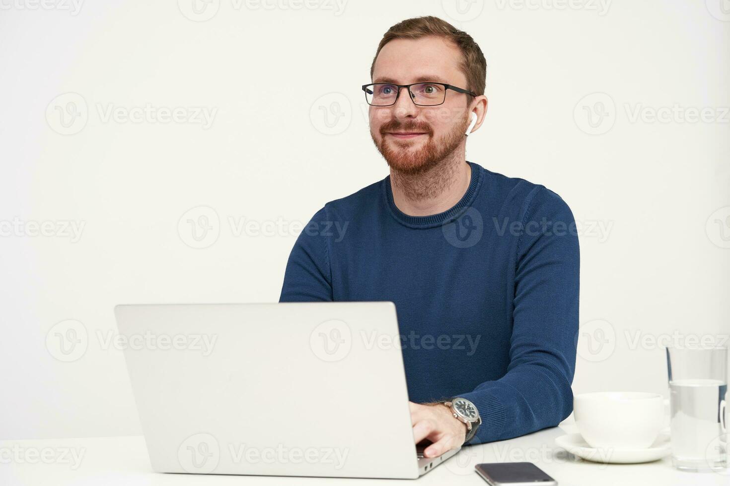 Horizontal shot of young pleased bearded fair-haired man looking positively ahead with lovely smile while keeping hands on keyboard, posing over white background photo