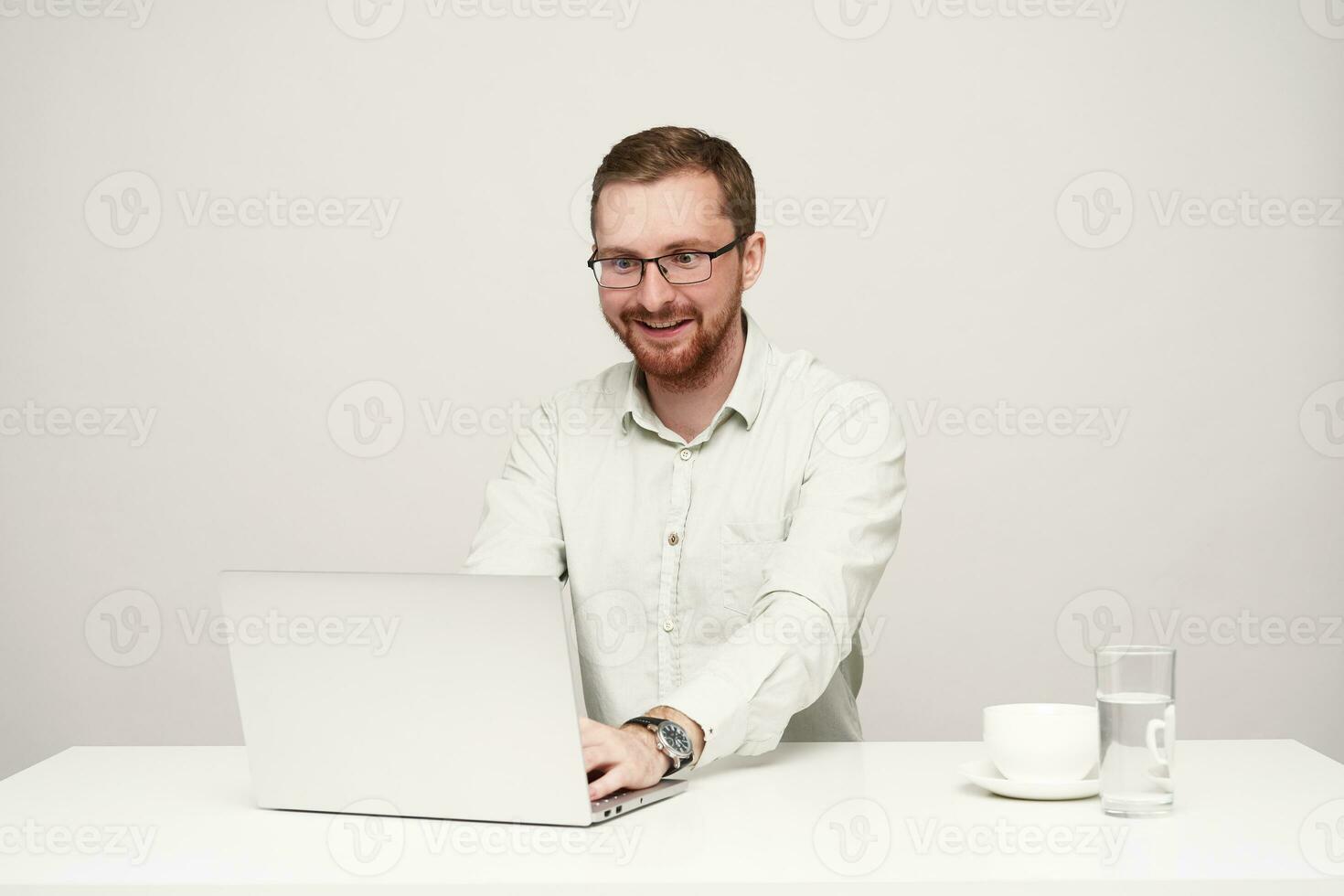 Surprised young pretty bearded fair-haired male in glasses looking excitedly at screen of his laptop while reading unexpected news, posing over white background photo