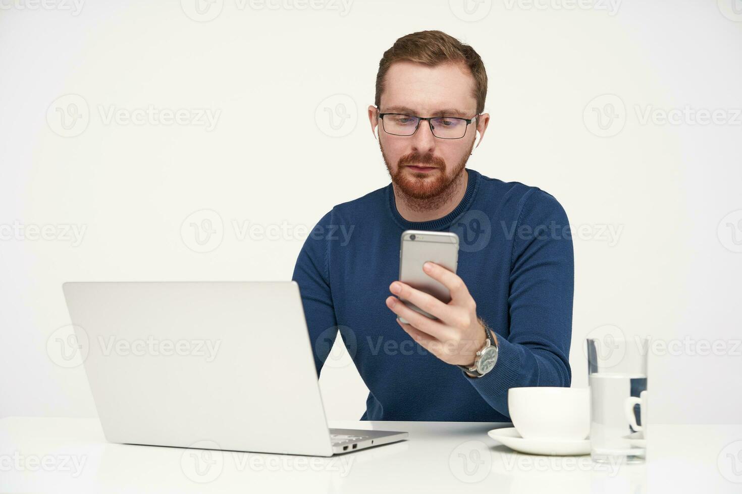 Studio photo of young fair-haired male in glasses keeping smartphone in raised hand and looking seriously on screen while reading message, posing over white background