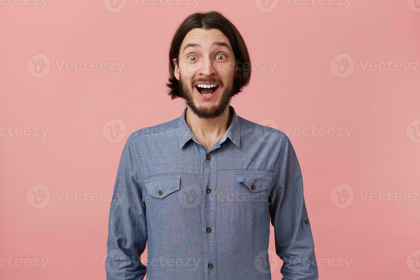 Photo of happy cheerful amazed bearded young man with long combed dark hair in denim shirt, heard the good news, was delighted, with open mouth, isolated over pink background.