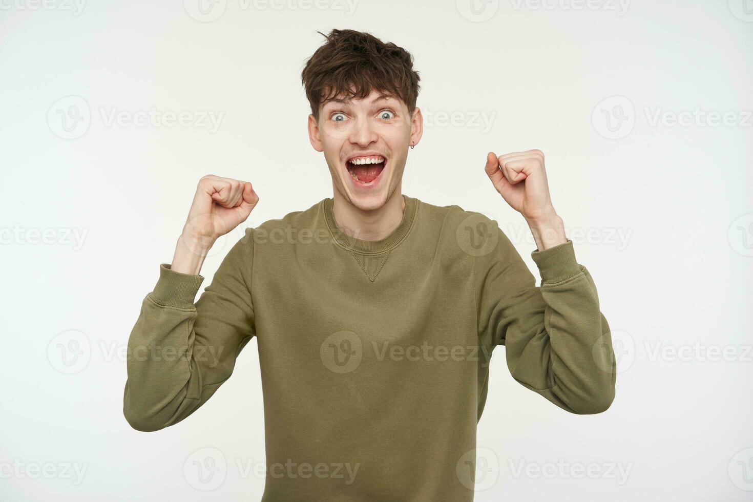 Teenage guy, happy looking man with brunette hair, piercing and bristle. Wearing khaki color sweater. Lifts up hes fists. Emotional concept. Watching at the camera isolated over white background photo
