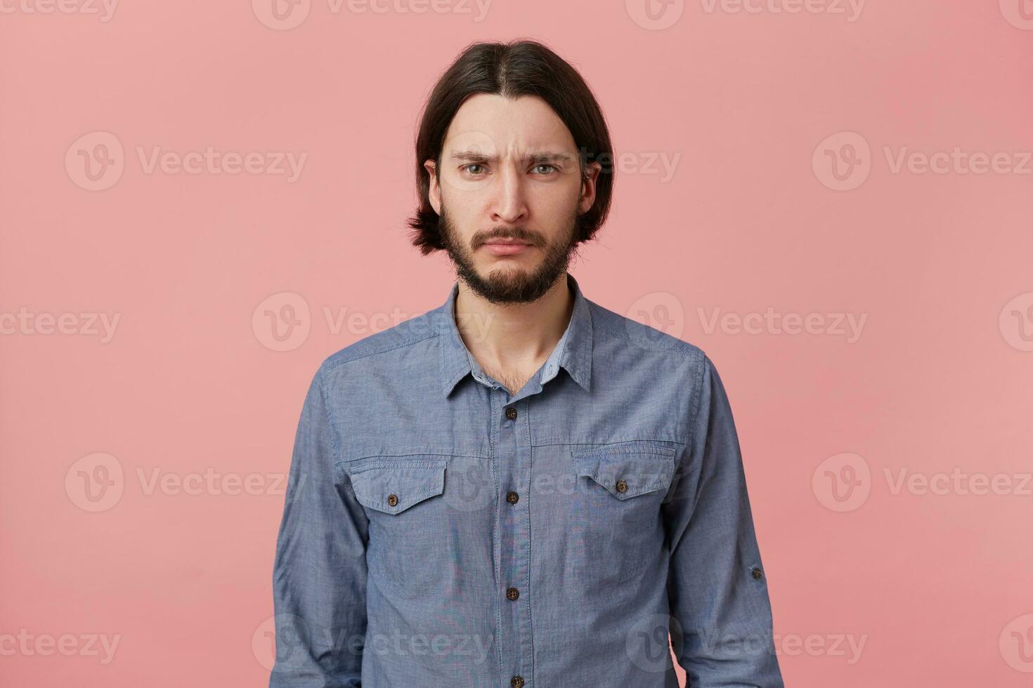 Photo of bearded guy with long dark hair looks sad upset hopeless, ready to cry, pouted his lips, isolated over pink background.