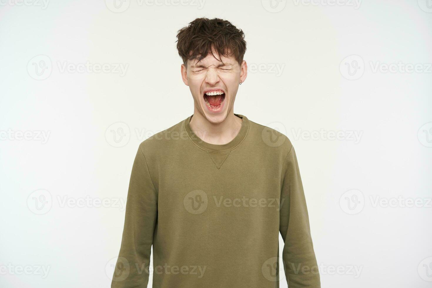 Cool looking male, handsome guy with brunette hair, piercing and bristle. Wearing khaki color sweater. Yelling hard with closed eyes, shout in anger. Stand isolated over white background photo