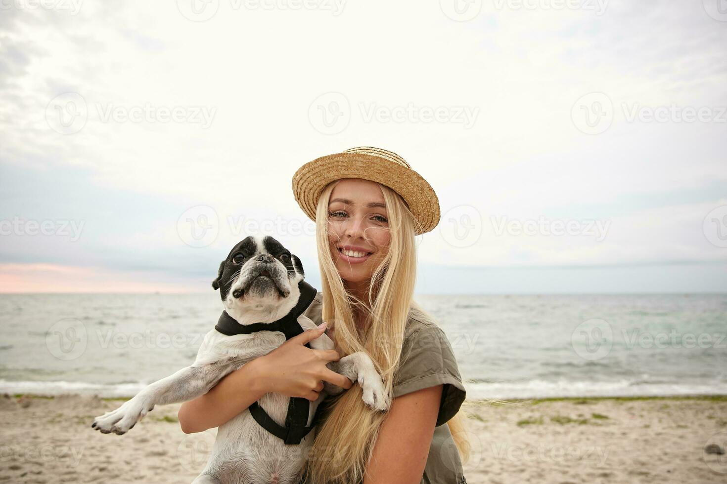 Photo of joyful pretty long haired female with blonde hair holding her funny dog on hands, looking in camera with cheerful smile while walking along beach on gray cloudy day