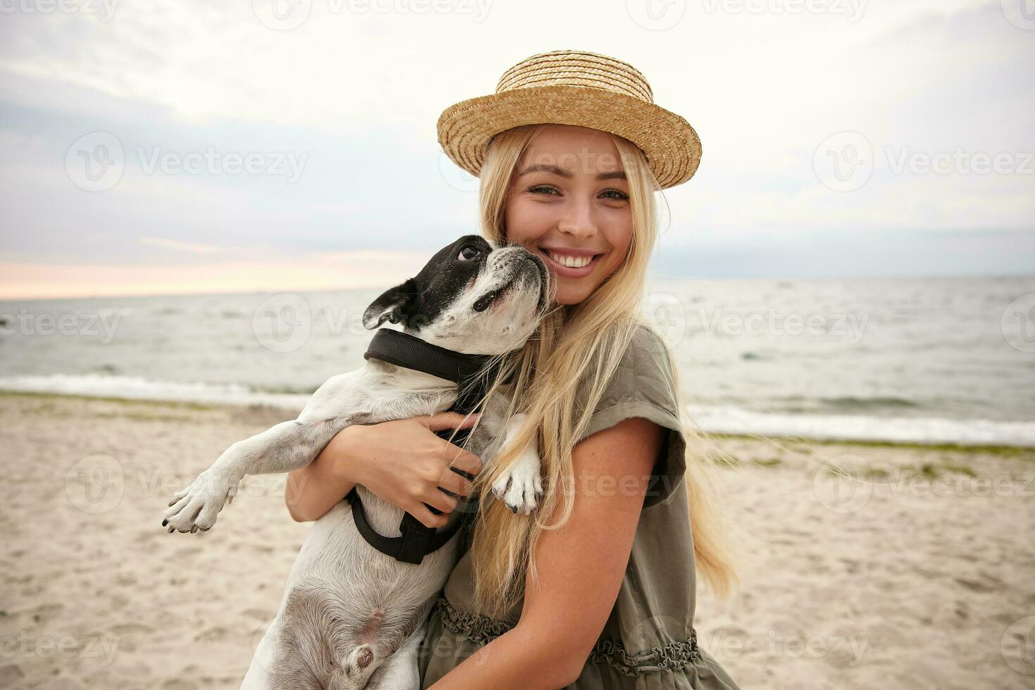 Portrait of happy young blonde female with long hair looking at camera with charming smile while keeping her beloved dog in hands, posing over seaside on gray cloudy day photo