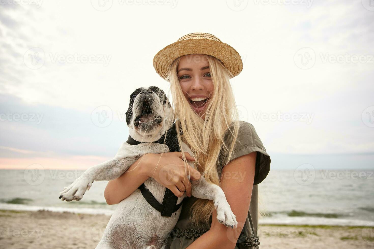 gracioso Disparo de hermosa joven mujer con largo rubia pelo vistiendo casual ropa, caminando a lo largo playa en nublado con su perro, mirando a cámara alegremente y sonriente extensamente foto
