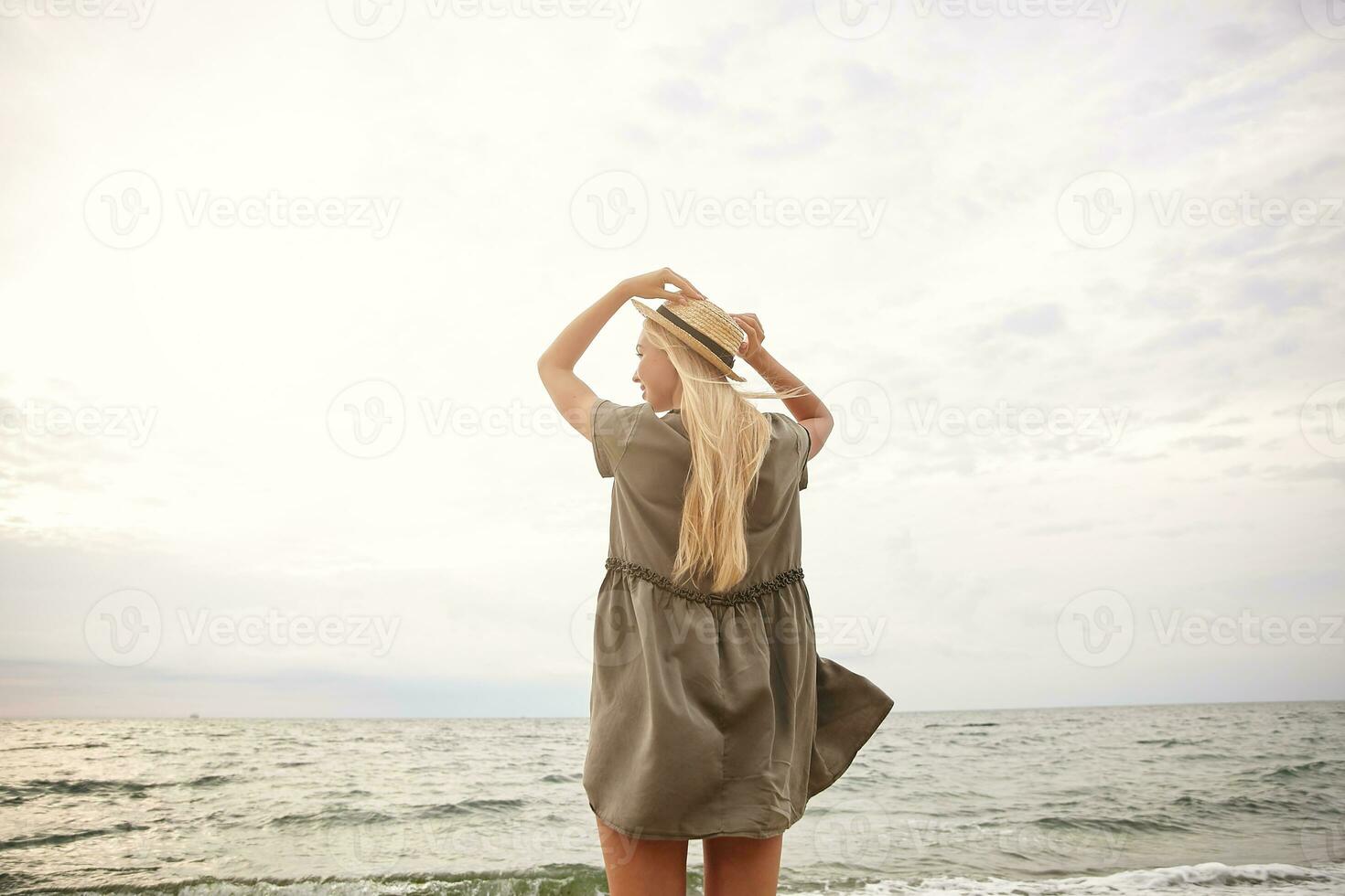 Bright outdoor photo of young slimwhite-headed female holding raised hands on her straw hat while enjoying the seaside view, wearing romantic green dress over beach background