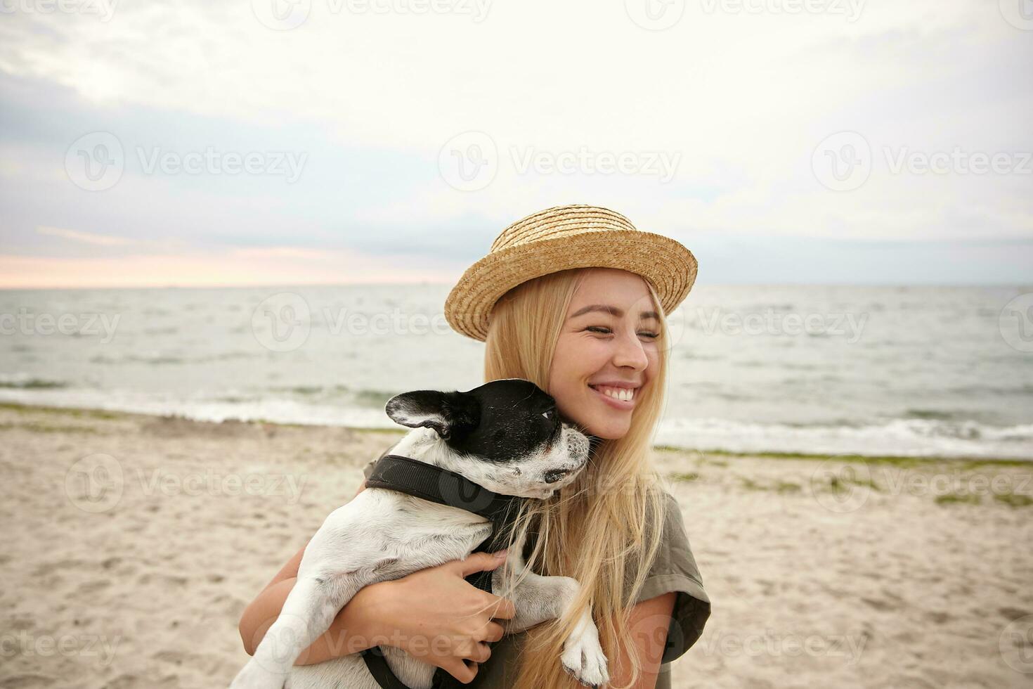 Outdoor photo of young pretty blonde with casual hairstyle standing over seaside and carrying her small dog on hands, smiling happily with closed eyes, wearing summer dress and boater hat