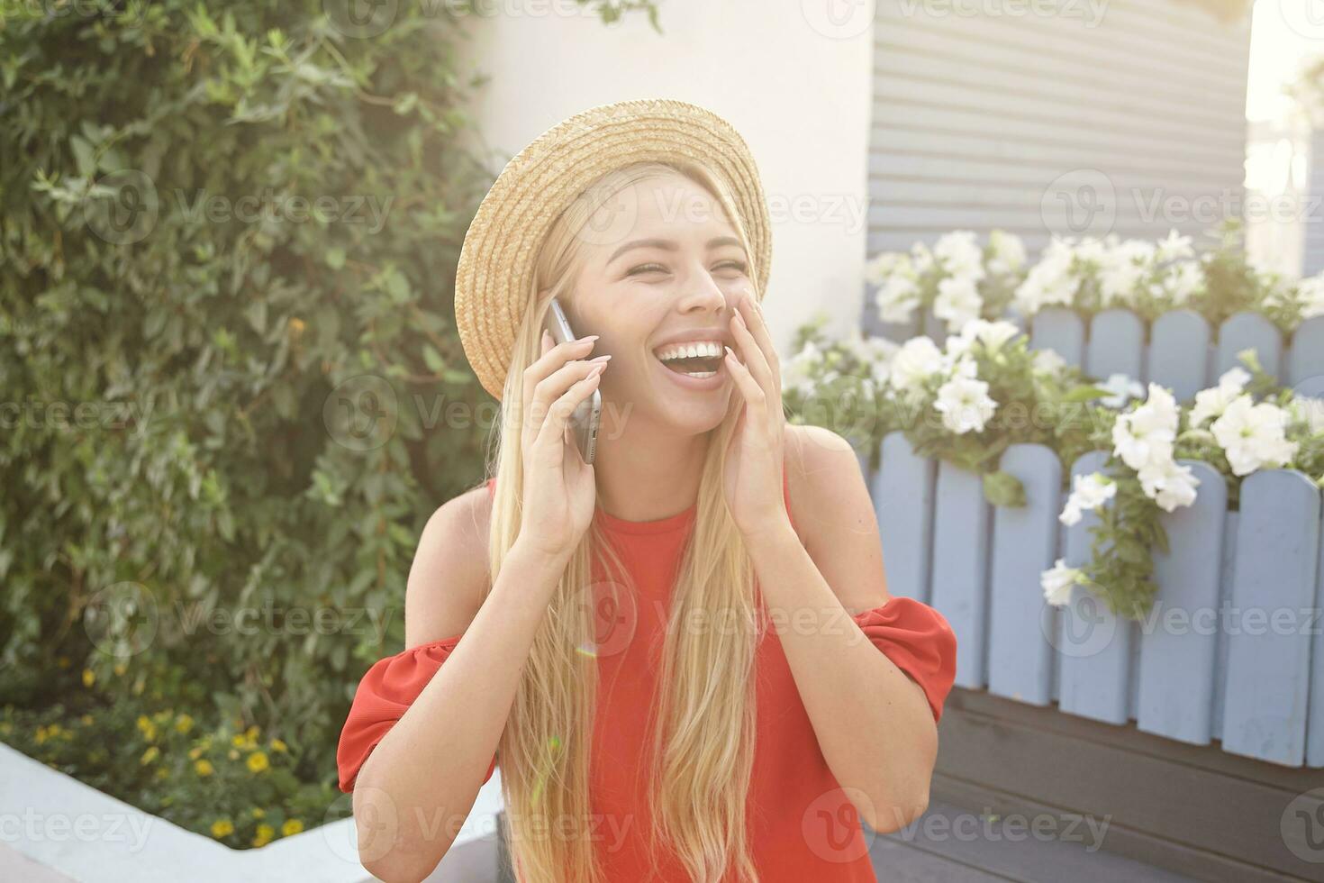 Happy young beautiful long haired blonde dressed in summer clothes covering her mouth with raised hand while laughing cheerfully during pleasant phone conversation photo