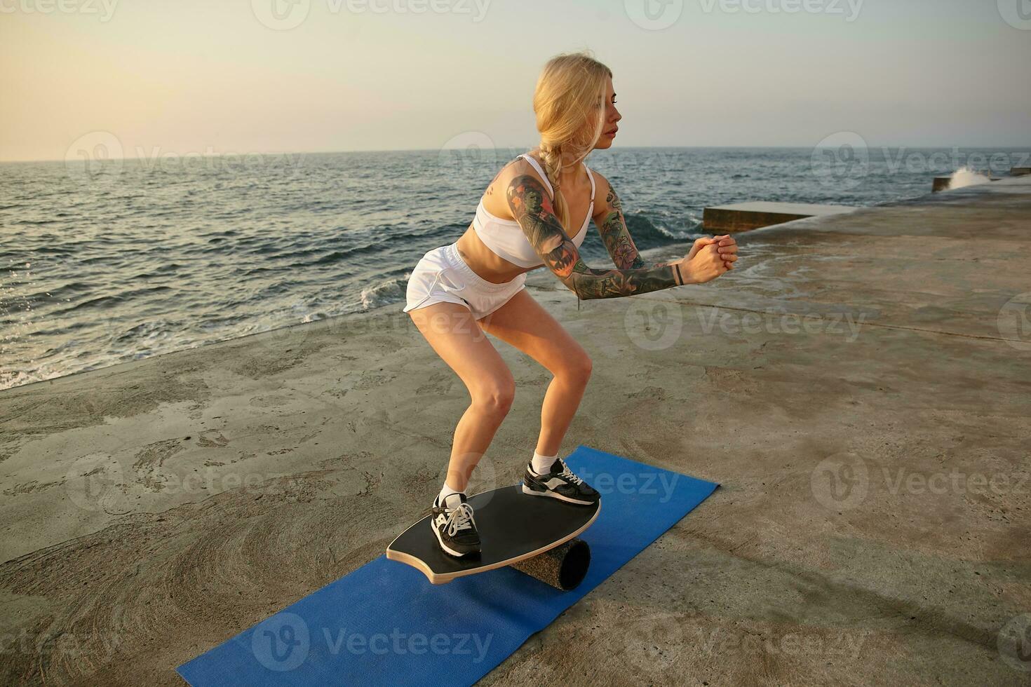 Outdoor photo of young sporty tattooed woman with long blonde hair combed in braid balancing on wooden board, raising folded hands and looking intently in front of herself, posing over promenade