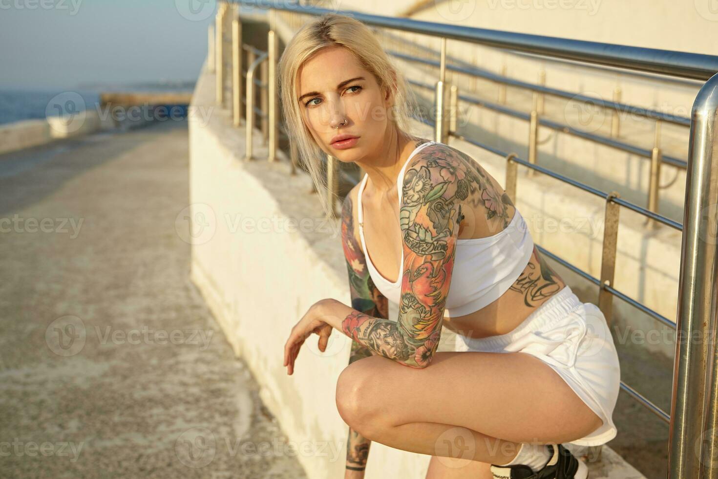 Attractive tattooed young blonde female with casual hairstyle posing over seaside promenade on early summer morning, wearing sporty clothes and sneakers, looking aside thoughtfully photo