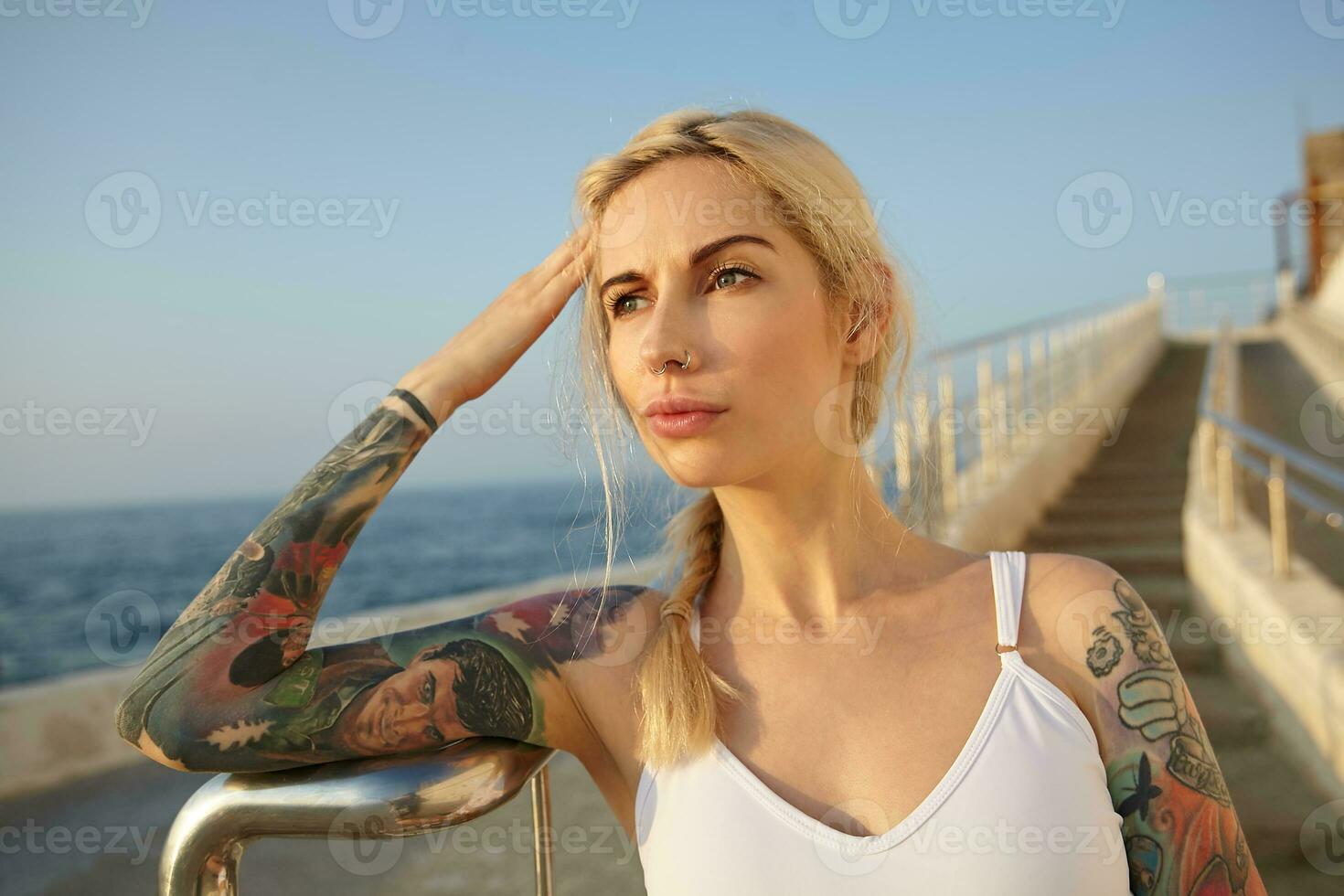 Outdoor close-up of beautiful blonde young female with tattooes meeting the dawn, looking aside with serious face and holding hand on her forehead, standing over seaside promenade photo