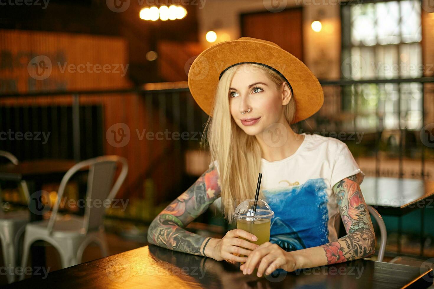 Beautiful young blonde female with tattooes wearing casual clothes, sitting at table in cafe and having cup of lemonade on lunch break, looking aside and smiling gently photo