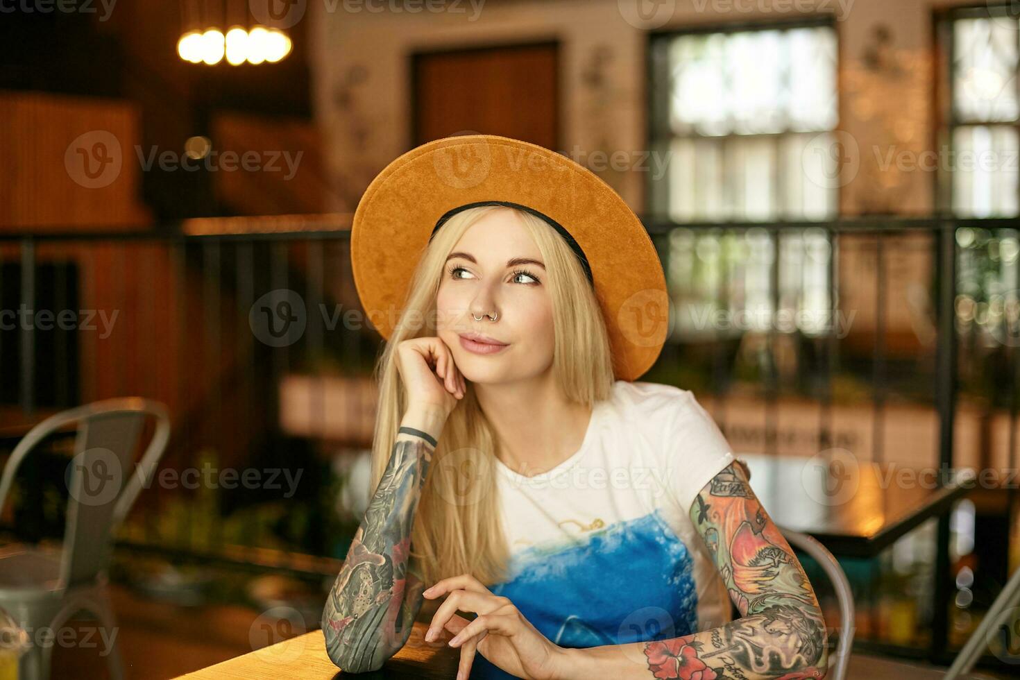 Photo of pretty young long haired woman with casual hairstyle sitting at table in cafe, looking aside positively while waiting for her order, wearing white and blue t-shirt and brown hat