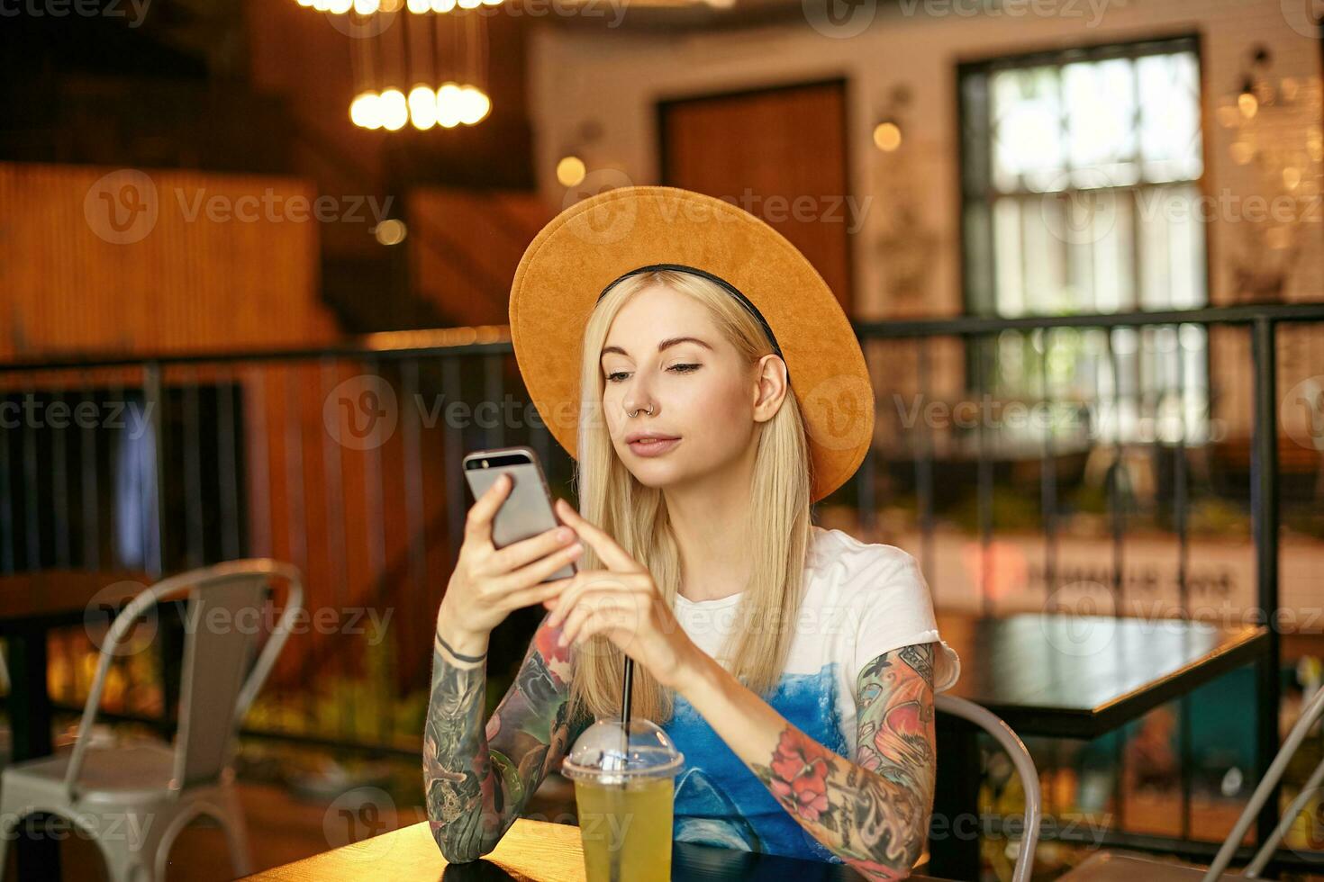 Indoor photo of pretty blonde long haired female with tattooes sitting at table in cafe and keeping mobile phone in hands, waiting for friends and drinking lemonade