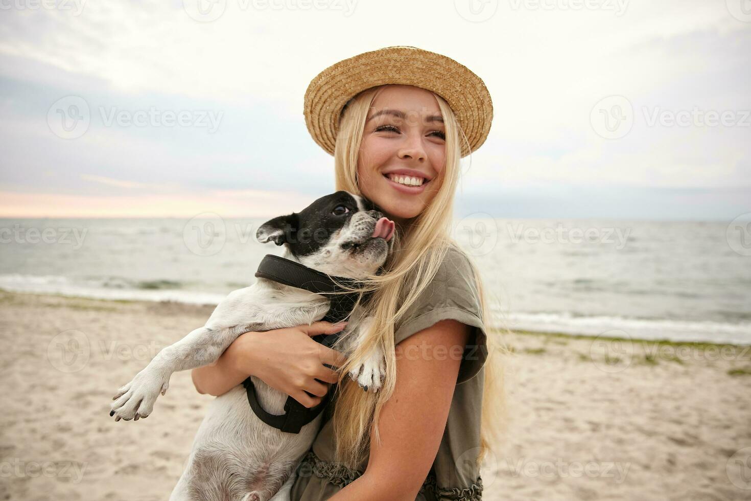 Joyful young pretty lady with blonde long hair posing over seaside on overcast with her french bulldog on hands, looking aside and smiling cheerfully, wearing casual clothes and straw hat photo