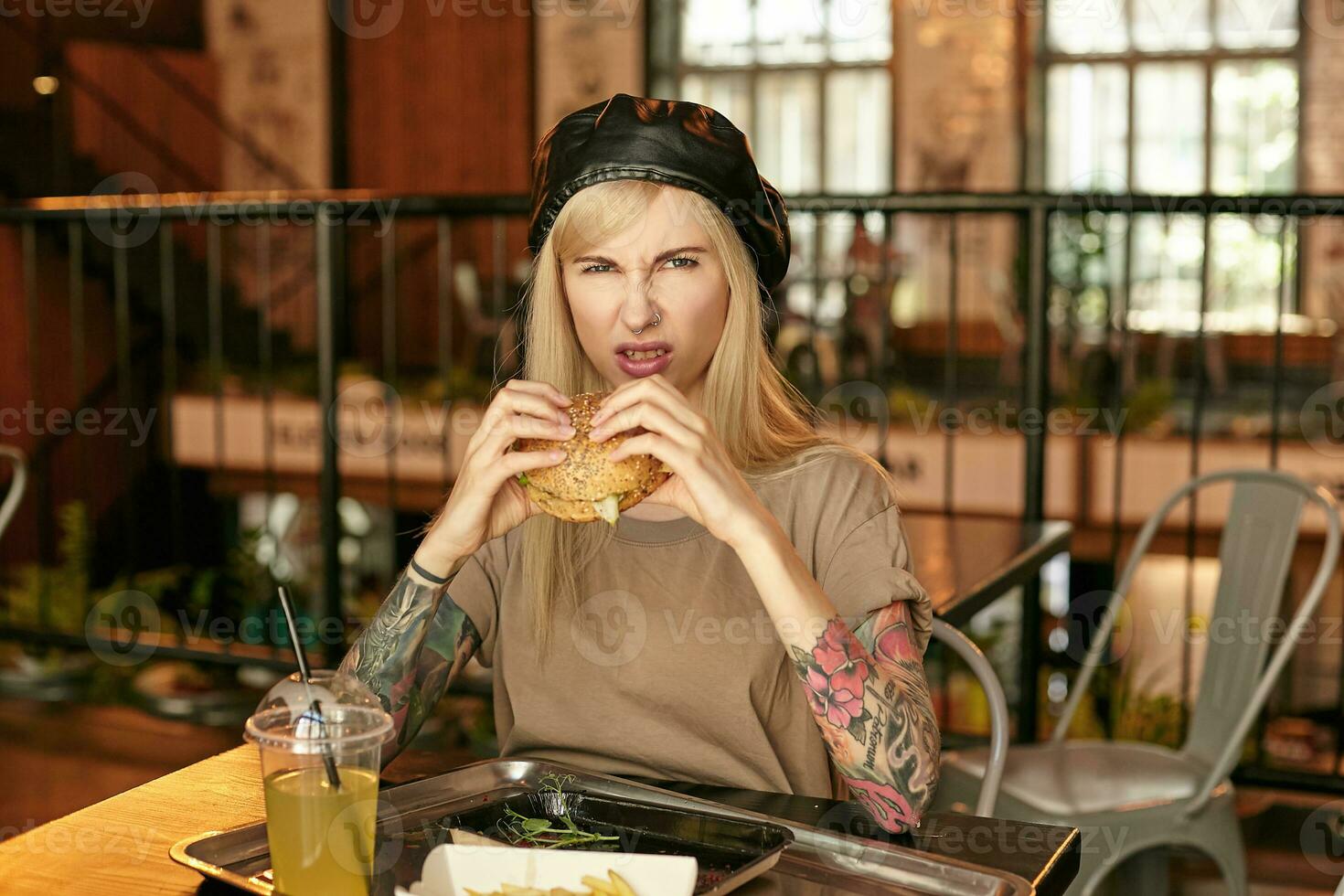 Indoor photo of unpleased tattooed blonde female posing over restaurant interior, trying hamburger and do not like it, wearing beige t-shirt and leather cap, looking at camera with pout