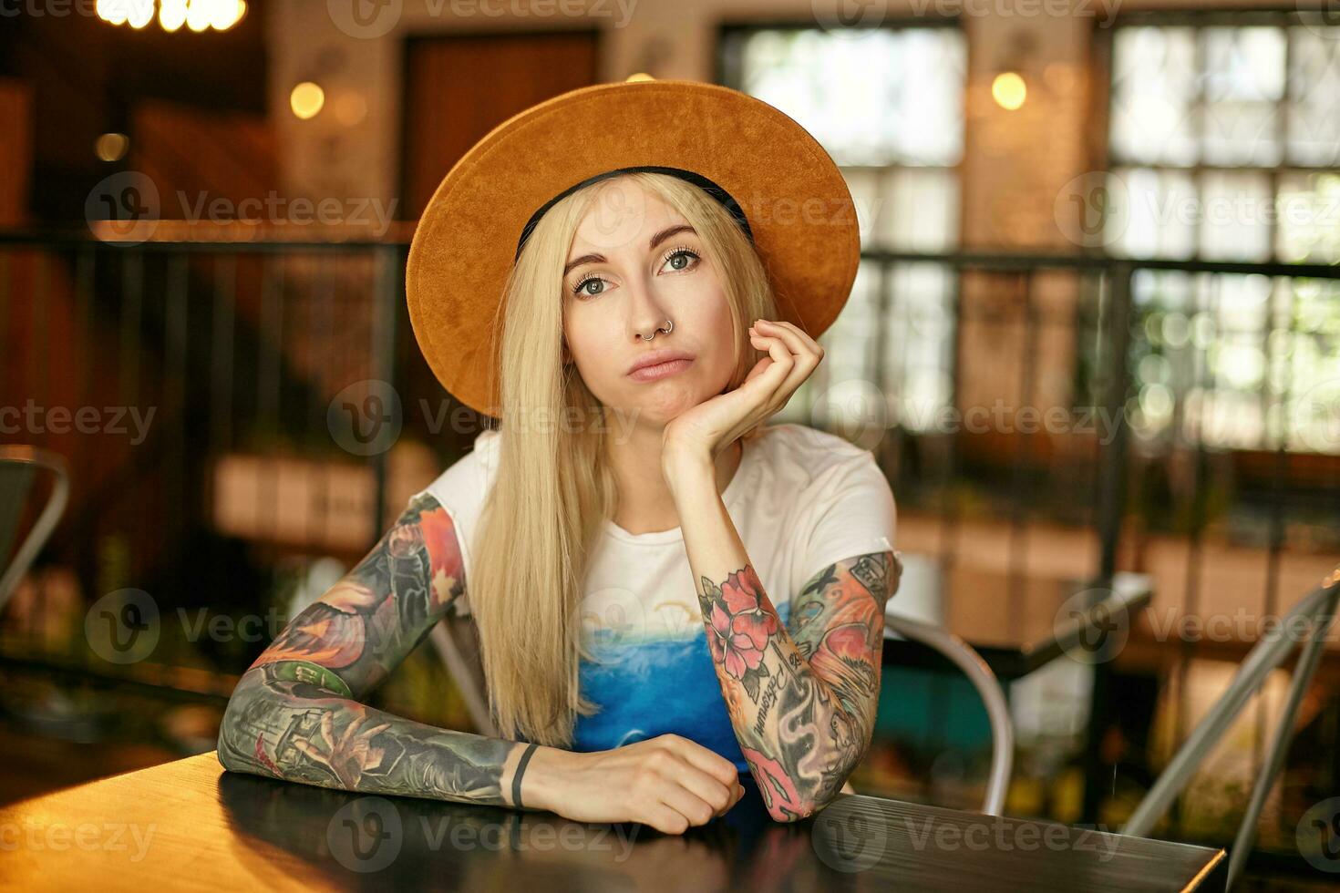 Indoor shot of puzzled blonde female with tattooes with posing over modern interior, sitting at table in cafe and leaning chin on raised hand, looking at camera with folded lips photo