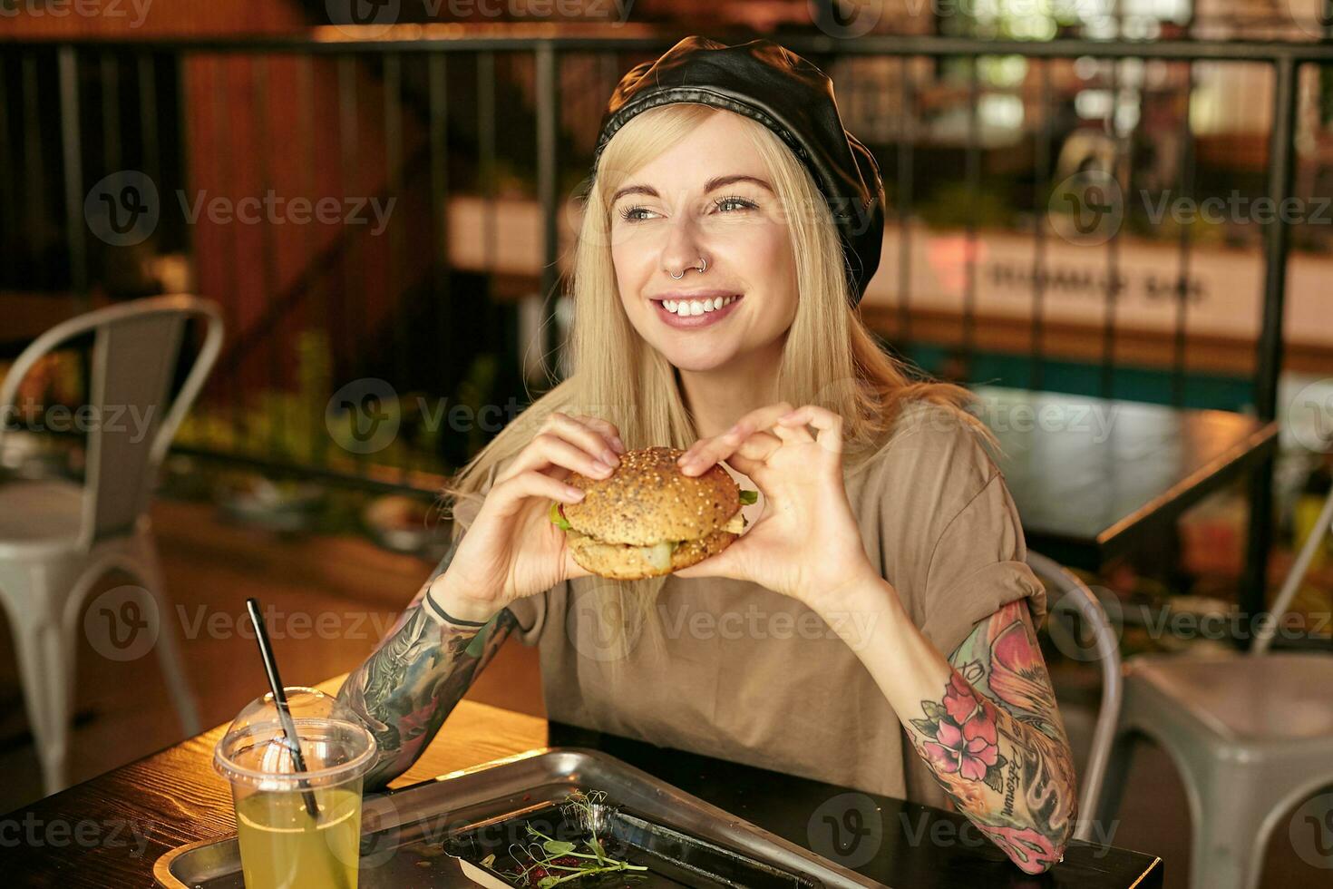 Horizontal shot of pretty tattooed blonde female in beige t-shirt and leather black beret posing over modern cafe interior, looking aside with wide charming smile and holding burger in hands photo