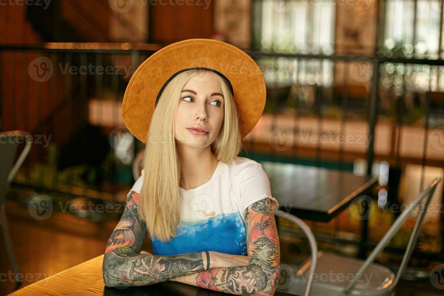 Indoor photo of attractive tattooed blonde woman in white and blue t-shirt and wide hat sitting over cafe interior and keeping folded hands on table, looking aside with light smile